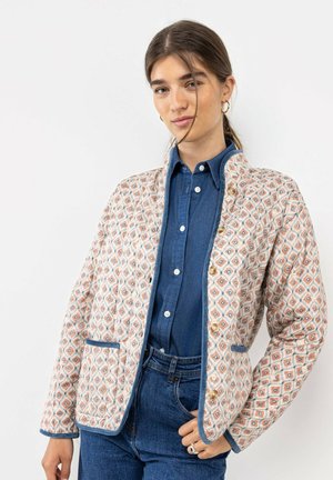 Joven mujer que lleva una chaqueta con botones y estampado sobre una camisa vaquera azul y vaqueros, de pie con una mano en el bolsillo sobre un fondo blanco.