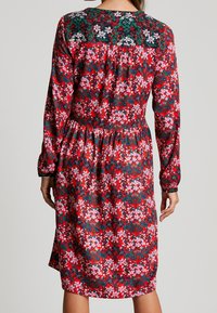 Czerwona sukienka w kwiaty z długimi rękawami, z wzorowanym gorsetem w ciemnozielonym i białym kolorze. Materiał jest gładki z marszczoną talią.