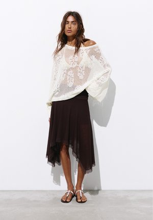 Femme portant un pull blanc en maille lâche sur un haut noir, associé à une jupe noire asymétrique et des sandales décorées de blanc, debout contre un mur blanc.