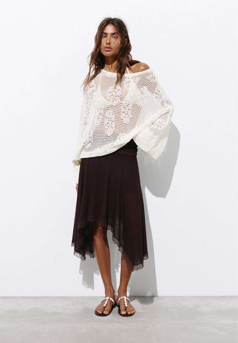 Femme portant un pull blanc en maille lâche sur un haut noir, associé à une jupe noire asymétrique et des sandales décorées de blanc, debout contre un mur blanc.