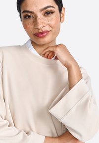 Jersey beige con cuello redondo y mangas amplias, hecho de una tela suave. Parte de una camisa blanca con cuello es visible debajo.