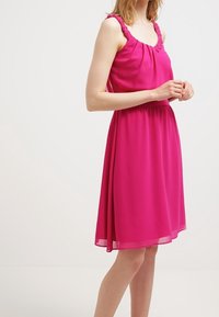 Robe sans manches fuchsia avec un décolleté froncé et des bretelles à volants. Tissu fluide tombant jusqu'au milieu du mollet, avec une taille cintrée.