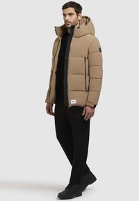 Beige gewatteerde jas met een capuchon, voorzien van een rits aan de voorkant en zwarte accenten. G gedragen over een zwart shirt en gecombineerd met zwarte broeken en laarzen.