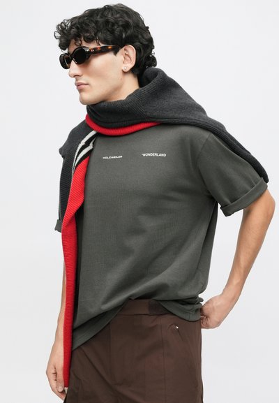 Olivno zelena majica s zavrnutimi rukavi, na kateri je bel napis. Temno siva ruta z rdečimi poudarki je spuščena čez ramena.