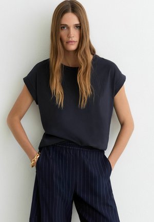 Mujer joven con cabello largo castaño claro, vestida con una camiseta negra de manga corta y pantalones azul oscuro con rayas finas, de pie con las manos en los bolsillos.