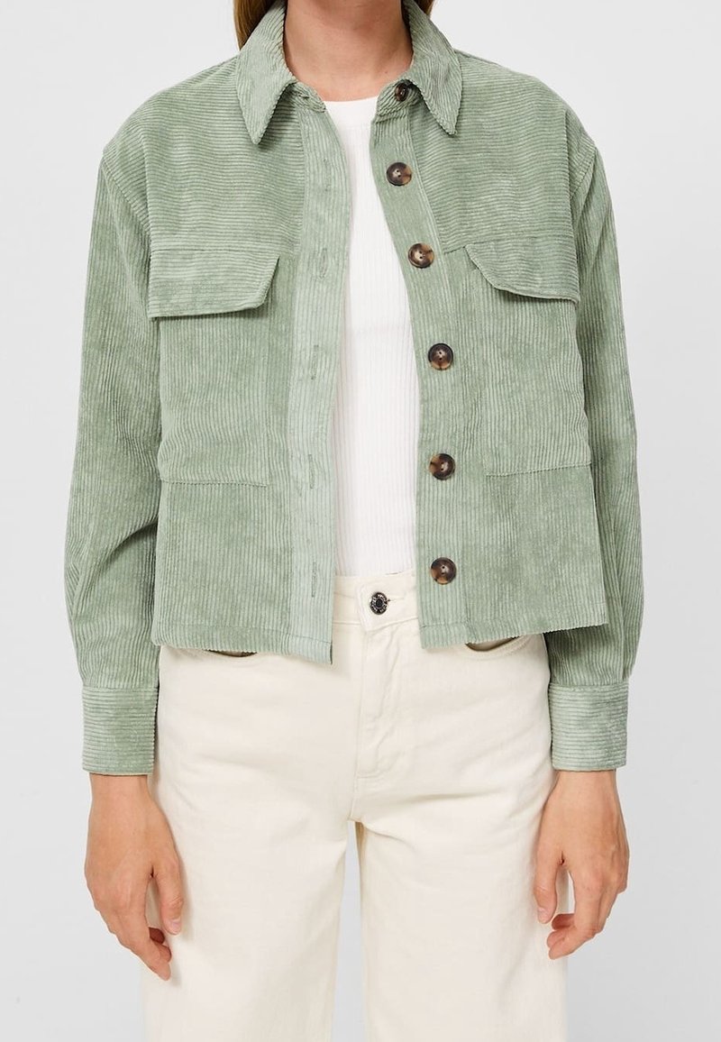 Femme portant une veste en velours côtelé vert clair à boutons avec deux poches poitrine à rabat, par-dessus un haut blanc et un pantalon crème.