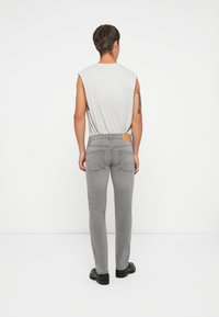 Jeans in denim grigio con una vestibilità slim, due tasche posteriori e un accento in pelle marrone chiaro sulla vita. Top senza maniche grigio. Scarpe nere.