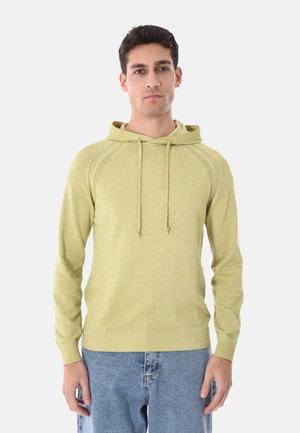 Giovane uomo con felpa con cappuccio verde chiaro e coulisse, indossa jeans blu, in piedi contro uno sfondo bianco uniforme.