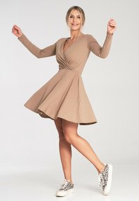 Beżowa sukienka z długim rękawem i rozkloszowanym dołem; wykonana z miękkiego materiału; zestawiona z wzorzystymi trampkami. Prosty dekolt i detal w pasie widoczne.