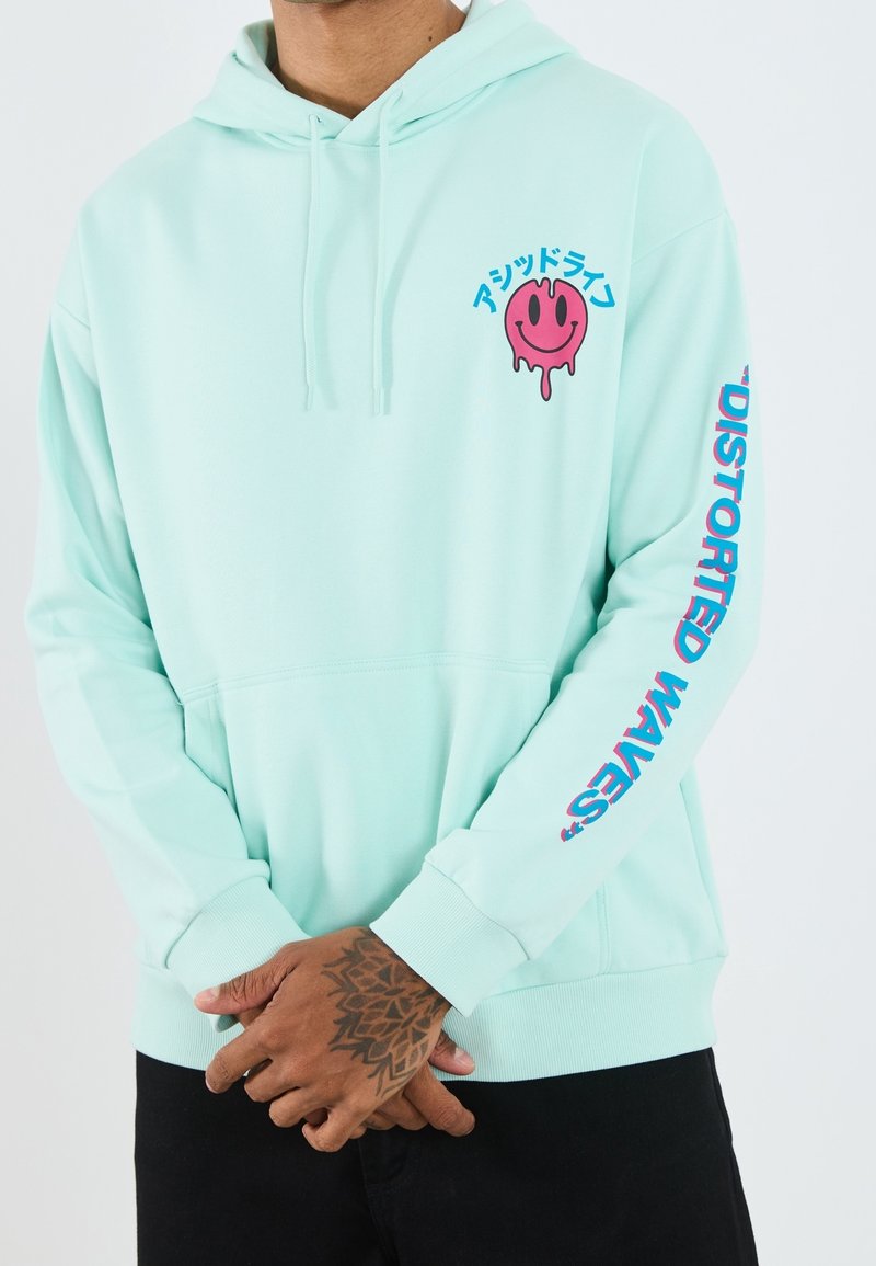 Homme en sweat à capuche vert menthe avec un visage souriant rose fondu et un texte japonais sur la poitrine, avec le texte "Distorted Waves" sur la manche droite.