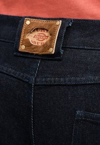 Κοντινή λήψη του πίσω μέρους σκούρων μπλε τζιν με καφέ δερμάτινο έμπλαστρο με υφή γούνας που δείχνει το λογότυπο της Dickies.