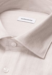 Camicia bianca con texture a spina di pesce, presenta un colletto classico, chiusura con bottoni e un'etichetta con la scritta "seidensticker."
