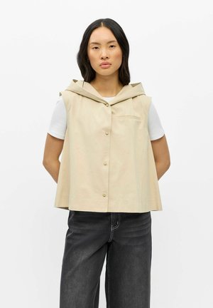 Kvinde iført beige ærmeløs hættetrøje vest over hvid kortærmet skjorte og sorte brede jeans, stående mod en hvid baggrund.