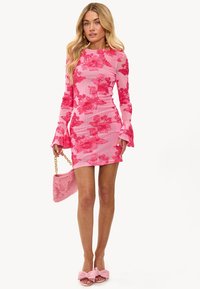 Vestido mini de estampado floral en tonos rosas con mangas largas y puños acampanados, que presenta un diseño fruncido. Combinado con un bolso de peluche rosa y sandalias decoradas con un lazo.