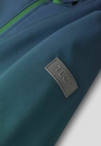 Tissu de veste bleu-vert au toucher lisse, avec une étiquette grise "TECH". Le design inclut un détail de fermeture éclair verte contrastante.