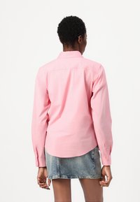 Camicia rosa a maniche lunghe con colletto standard, spallina posteriore e polsini con bottone. Il tessuto appare liscio con una leggera lucentezza.