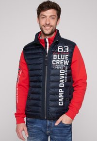 Gilet imbottito blu navy con zip frontale, grafiche di testo bianche e una maglietta rossa a maniche lunghe sotto. Presenta un colletto alto e una tasca laterale.