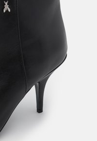 Bota de tacón alto de cuero negro con punta afilada, que presenta una textura suave y un acento metálico de abeja cerca del tobillo. Tacón de aguja delgada.