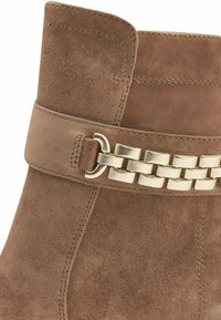 Botín de ante marrón que presenta una amplia banda decorativa con un acento de cadena de tono dorado, textura suave y costuras reforzadas.