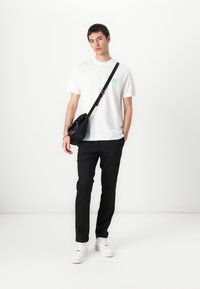 Valkoinen puuvillapaita vihreällä logolla, mustat housut, musta olkalaukku ja valkoiset tennarit. Yksinkertainen, moderni malli, jossa on kontrastivärejä.