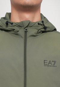 Oliven grønn hettejakke med glidelås. Har et teksturert stoff, justerbare snorer og en trykt EA7-logo på brystet.