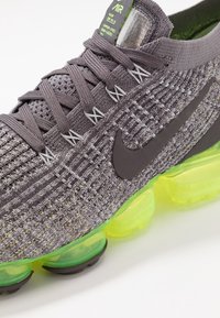 Sneaker Nike Air z szarym, dzianinowym wierzchem z czarnymi akcentami, zieloną podeszwą w kształcie bańki i charakterystycznym teksturowanym wzorem.