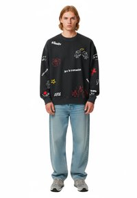 Felpa nera con grafiche colorate e testo. Abbinata a jeans azzurri chiari e sneakers grigie. Taglio casual e stile rilassato.