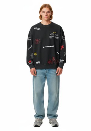 Felpa nera con grafiche colorate e testo. Abbinata a jeans azzurri chiari e sneakers grigie. Taglio casual e stile rilassato.