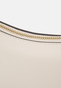 Sac en cuir beige avec une texture lisse et une forme incurvée. Dispose d'une fermeture éclair dorée et d'un contour sombre contrastant.