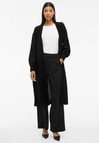 Cardigan noir en maille avec un motif texturé, à décolleté ouvert et à manches longues. Associé à un pantalon noir taille haute et des chaussures à bout pointu.