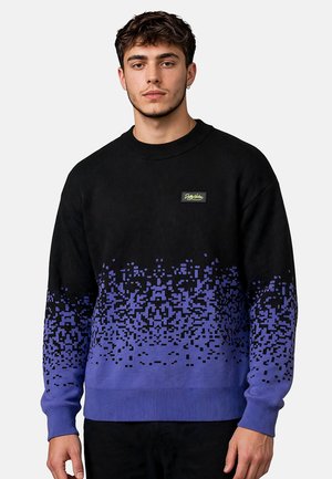 Joven vestido con un suéter negro y morado con patrón de píxeles y un pequeño logo rectangular en el pecho, de pie frente a un fondo liso.