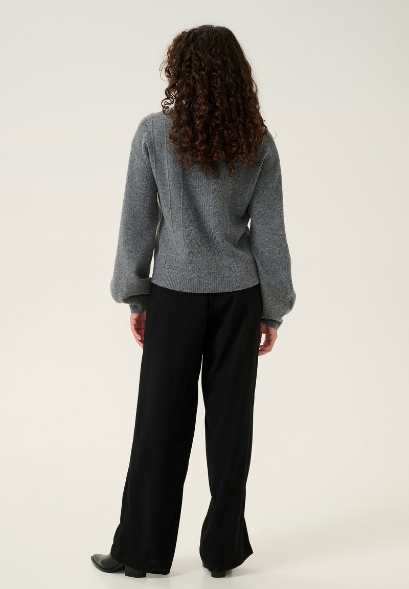 Grauer gestrickter Pullover mit einem strukturierten Muster, Glockenärmeln, kombiniert mit schwarzen weit geschnittenen Hosen und Ankle Boots, von hinten betrachtet.