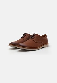 Chaussures en cuir marron avec une texture lisse, bout arrondi et semelle en caoutchouc plus claire contrastante. Présente des lacets et des accents cousus sur les côtés.