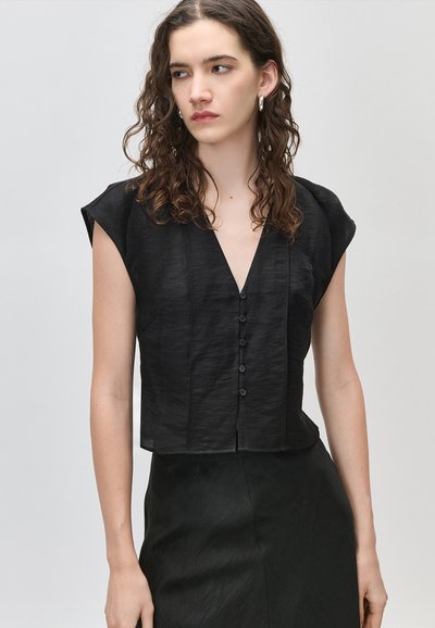 Νεαρή γυναίκα με σγουρά μαλλιά που φοράει μαύρο αμάνικο τοπ με λαιμόκοψη σε σχήμα V και κουμπιά, καθώς και matching φούστα, μπροστά σε απλό γκρι φόντο.