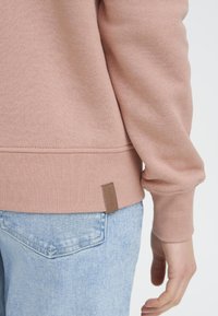 Różowa bawełniana bluza z prążkowanymi mankietami, z brązową skórzaną metką. Noszona z jasnoniebieskimi jeansami denimowymi. Miękka faktura i swobodny krój.