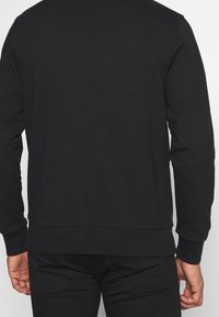 Sudadera negra con una textura suave, puños y dobladillo acanalados, diseño de cuello redondo y detalles mínimos, combinada con pantalones negros.