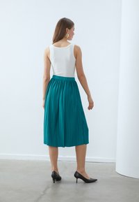 Teal, plissert midiskjørt med en tettsittende midje, kombinert med en hvit tanktopp og svarte hæler, sett fra bak.