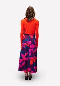 Une femme avec des cheveux bruns ondulés porte un haut à manches longues orange vif et une jupe maxi florale colorée taille haute avec des talons noirs.