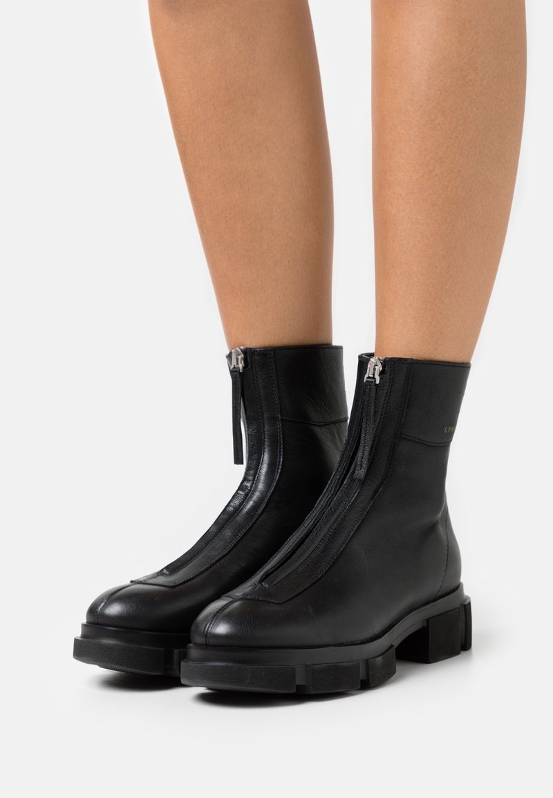 Bottes chevilles en cuir noir avec zips frontaux et semelles épaisses portées sur jambes nues sur fond blanc uni.