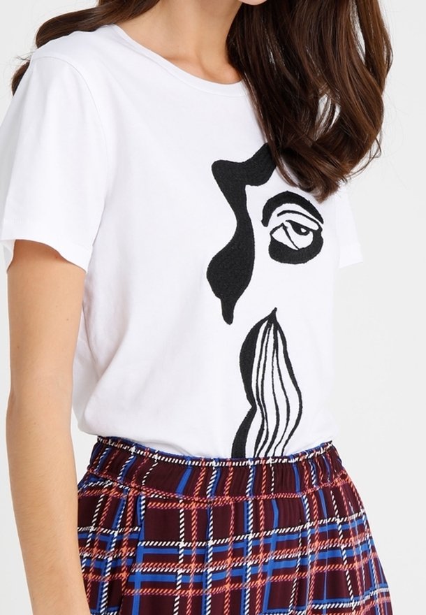 T-shirt en coton blanc avec un motif abstrait de visage noir. Associé à une jupe à carreaux bordeaux, bleue et blanche avec une finition texturée.