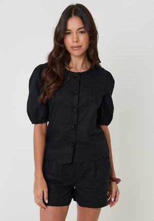 Kvinde med langt, bølget hår iført en sort knapbluse med ballonærmer og matchende sorte shorts foran en ensfarvet baggrund.