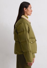 Olivgrüner Pufferjacke mit boxy Silhouette, ausgestattet mit einem hellbeigen Kragen und einer Fronttasche mit Druckknopf. Glatte Textur.