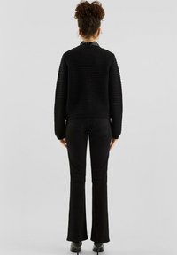 Frau mit lockigem Haar in einem schwarzen strukturierten Pullover, schwarzen Schlaghosen und schwarzen High Heels, die mit dem Rücken zu einer einfarbigen Hintergrund steht.