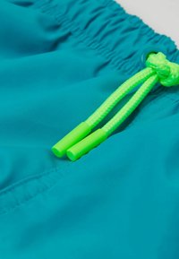 Tyrkysové plavkové šortky s elastickým pasem, se světlými zelenými stahovacími šňůrkami a hladkým, lehkým materiálem. Žádné viditelné vzory.