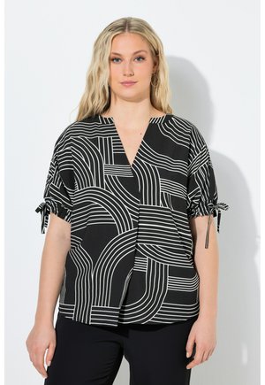 Mujer rubia que lleva una blusa de manga corta con patrón geométrico en blanco y negro y detalles de lazo en las mangas, de pie frente a una pared blanca.