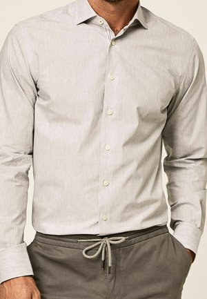 Homme portant une chemise beige clair à manches longues boutonnée, rentrée dans un pantalon gris avec une taille à cordon noué. Mains dans les poches.