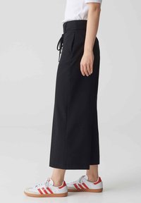 Zwarte wijde broek met een hoge taille, voorzien van een trekkoord en textured stof, gecombineerd met witte sneakers met rode accenten.