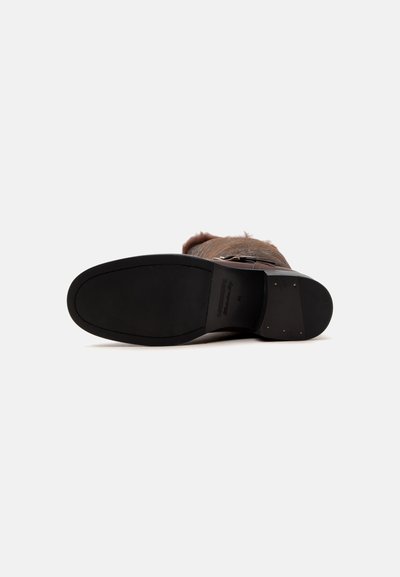 Vue de dessous d'une botte en cuir marron avec une semelle noire lisse et un petit talon clouté sur un fond uni.