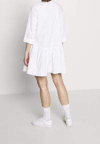 Abito bianco a maniche corte con gonna a ruota, realizzato in cotone leggero. Design semplice con dettaglio arricciato in vita. Abbinato a sneaker bianche.