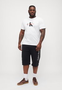 Hvit bomull t-skjorte med Calvin Klein-logo, kombinert med sorte knelengde shorts med hvit tekst på siden. Brune sneakers med hvite sokker.